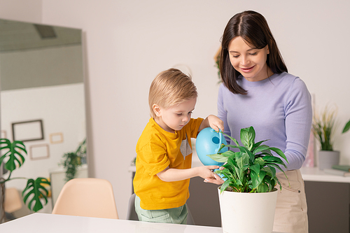 Smiling young mother assisting son to water domestic plant using watering can at home