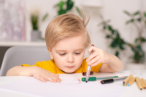 Concentrated cute blond-haired boy sitting at table and drawing on paper with colorful felt-tip pens