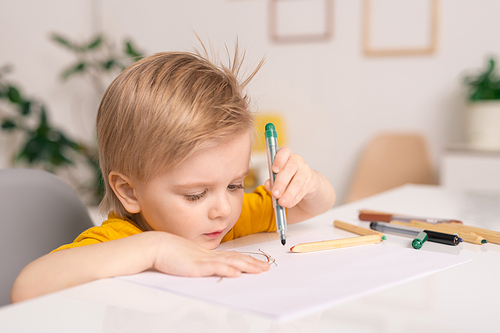 Cute blond little boy with green crayon or highlighter over paper drawing picture by desk while staying at home on quarantine