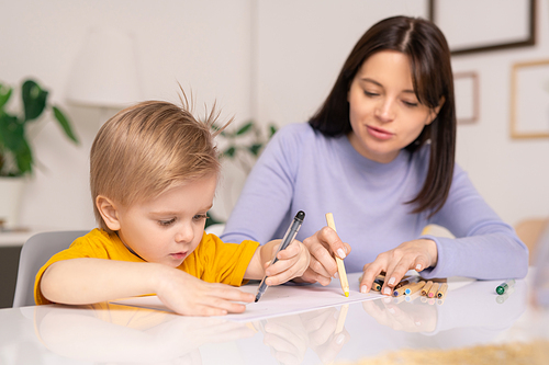 Careful young mother sitting at table and drawing on paper with little son