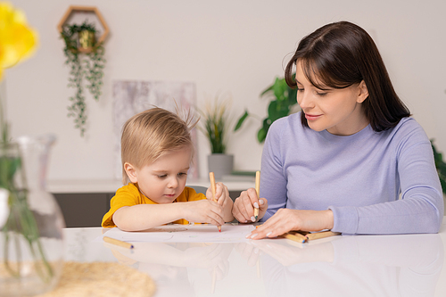 Pretty young mother and her cute little son drawing pictures with colorful crayons while staying at home during period of quarantine