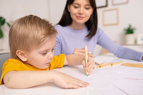 Close-up of concentrated cute boy sitting at table and using pencils for drawing under control of mother