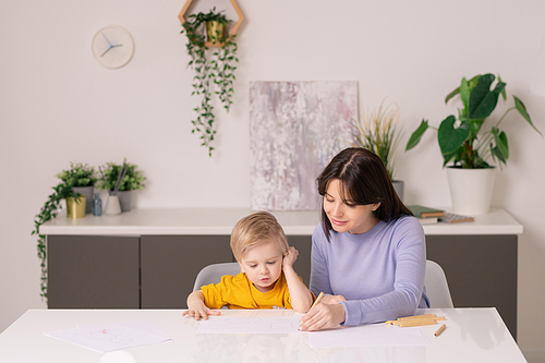 Young casual woman helping her cute little son to draw picture on paper with colorful crayons while both sitting by table in living-room