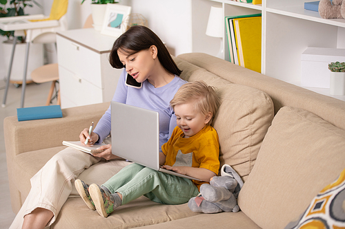 Happy cute little boy playing with laptop while sitting on couch next to his young mother talking on smartphone and making notes in notepad