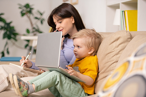 Curious little boy sitting on sofa and watching cartoon on laptop while mother answering phone call