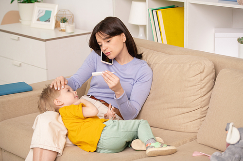 Worried young mother sitting on sofa and checking temperature of son while calling doctor by phone