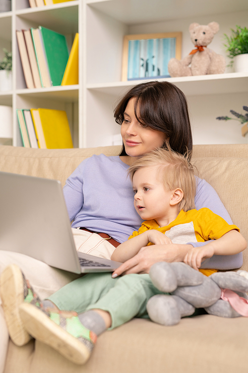 Blond-haired boy embraced by mom sitting on sofa and watching curious cartoon on laptop at home