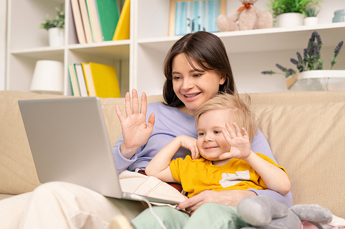 Cheerful young mother and son sitting on sofa and waving hands while talking to father via video chat