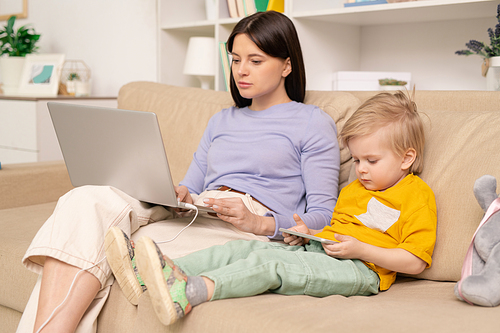 Young female in casualwear working remotely in front of laptop while sitting on couch next to her cute little son watching cartoon in smartphone