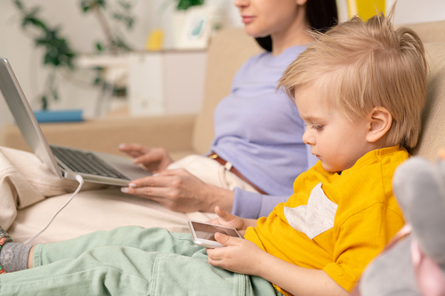 Close-up of serious concentrated kid boy sitting on sofa and using gadget while mother working with laptop