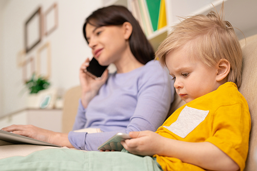 Cute blond little boy with smartphone watching online cartoon while his mother talking on the phone and working remotely on quarantine