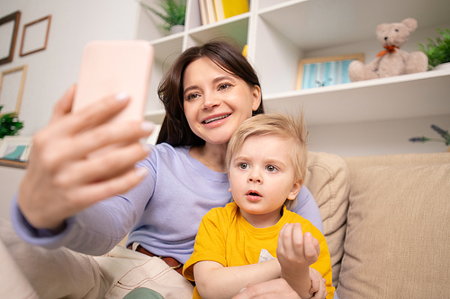 young smiling woman and her adorable son sitting on couch and  in smartphone while making selfie in home