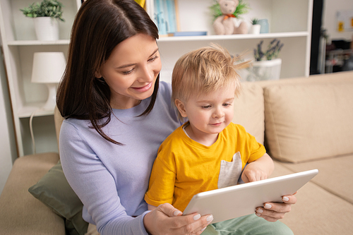 Happy young mother and cute little son looking through photos or watching video in digital tablet while relaxing on couch during quarantine