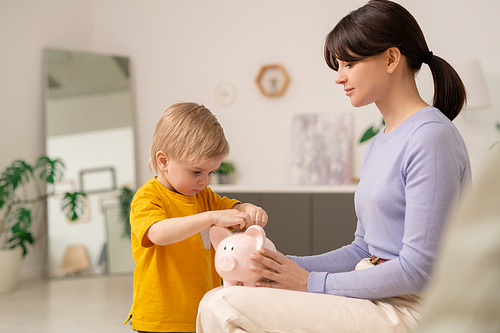 Young mother holding piggy bank while son putting coin into it, mother teaching son to save money