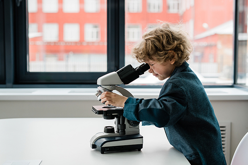 Clever schoolboy or biologist in casualwear bending over desk while looking in microscope against laboratory window