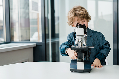 Clever cute schoolboy or biologist standing by desk while looking in microscope in classroom or laboratory