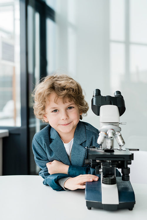 Cute little successful scientist looking at you while standing by microscope in classroom or laboratory