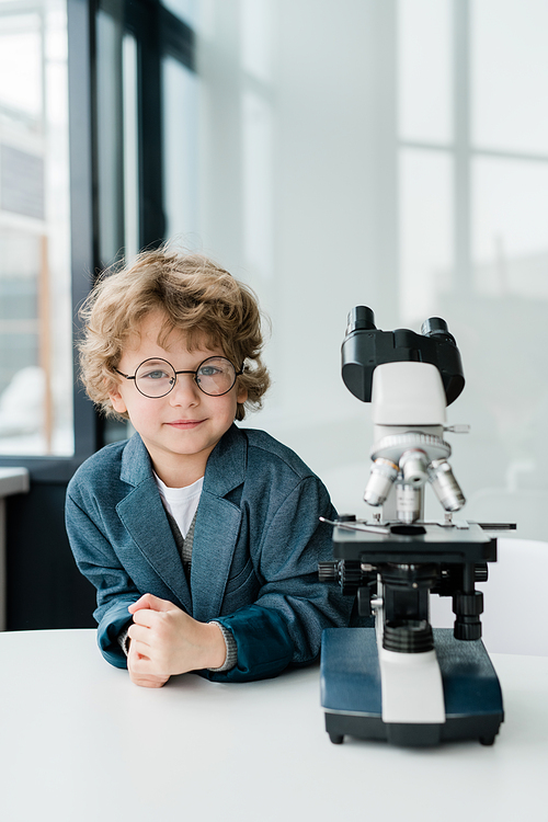 Cute little scientist in eyeglasses and formalwear looking at you while standing by microscope at workplace