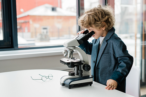 Clever schoolboy with curly hair bending over desk while looking in microscope during work in laboratory