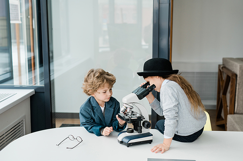 Two clever elementary schoolkids studying chemistry with microscope while working in group by desk at lesson