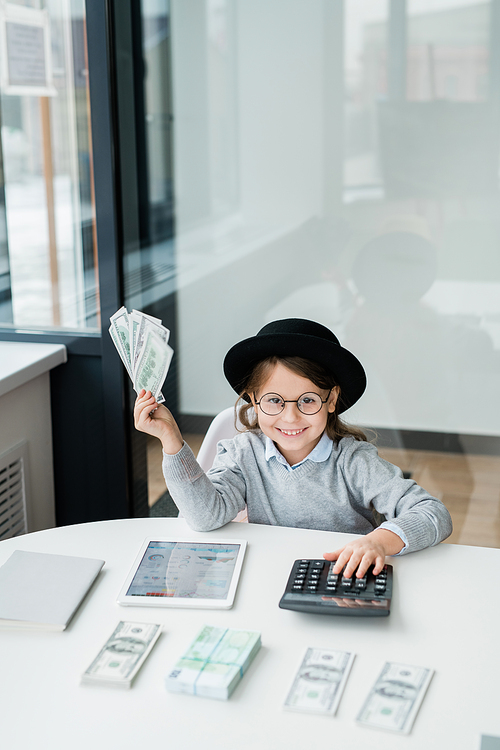 Little cheerful accountant in hat and casualwear looking at you while holding bunch of dollar banknotes over desk