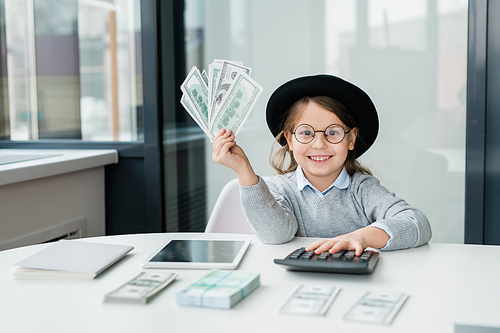 Little accountant with toothy smile counting your salary while holding bunch of dollar banknotes over desk