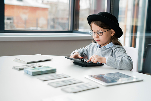 Little serious accountant in hat and eyeglasses using calculator while counting salary of office managers by desk
