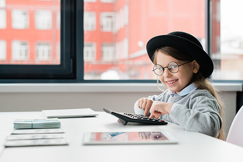 Little smiling accountant in hat and eyeglasses looking at you while counting salary of office managers by desk