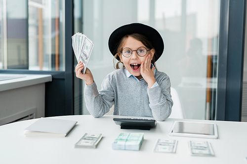 Amazed little accountant in casualwear looking at you while holding bunch of dollar banknotes over desk and touching her face