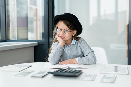 Little smiling accountant in eyeglasses looking at you while counting salary by workplace in office