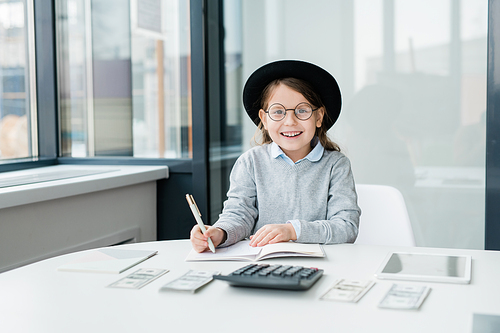 Cheerful little girl in hat and eyeglasses looking at you while counting money and making notes in copybook