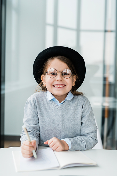Adorable and cheerful little girl in hat and eyeglasses looking at you while making notes in copybook