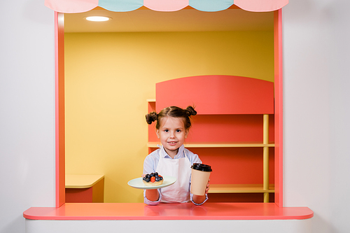 Cute little girl holding glass of drink and plate with berry cake or dessert while passing it to guests of cafeteria