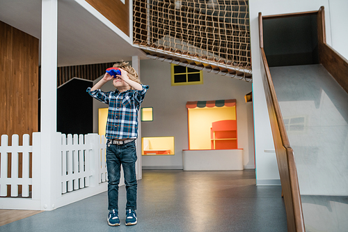 Cute little boy in casualwear looking through binoculars while standing on play area on background of slide