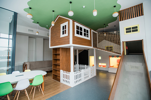 Play area of contemporary children leisure center with slide, small house, row of armchairs and table with chairs around