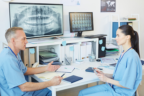 Side view portrait of two modern doctors wearing blue uniform discussing case while sitting at workplace in dental clinic, copy space