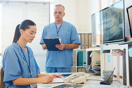 Portrait of two adult doctors wearing blue uniforms writing on clipboards while working at desk in clinic interior, copy space