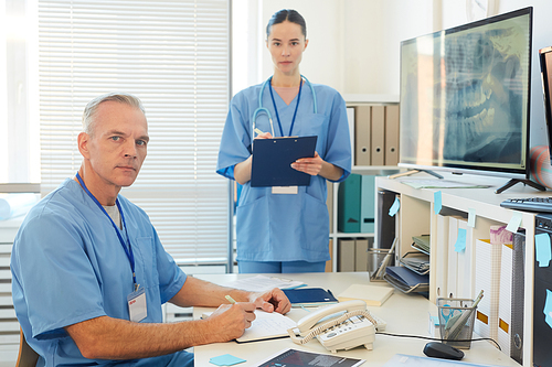 Portrait of two adult doctors wearing blue uniforms  while working at desk in modern clinic interior, copy space