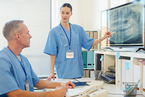 Waist up portrait of female medic pointing at x-ray image while talking to mature doctor sitting at desk in clinic, copy space