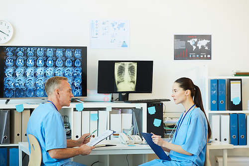 Side view portrait of two adult medics talking while sitting at workplace in modern clinic interior with CT scans in background, copy space