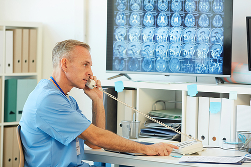 Side view portrait of mature doctor speaking by phone while sitting at desk in hospital interior with CT scans in background, copy space