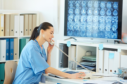 Side view portrait of young female medic speaking by phone while sitting at desk in hospital interior with CT scans in background, copy space