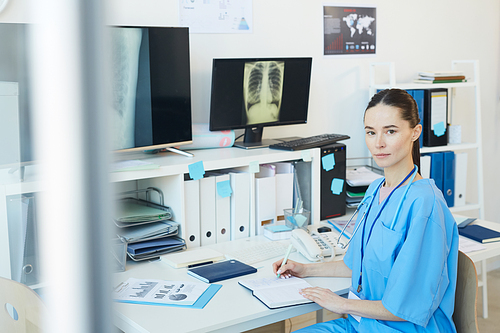 Waist up portrait of young female doctor  while posing at workplace with chest x-ray scans in background, copy space