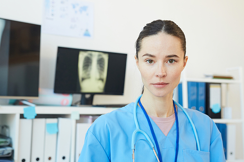Head and shoulders portrait of young female doctor  while posing at workplace with chest x-ray scans in background, copy space
