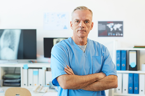 Waist up portrait of confident mature doctor  while standing with arms crossed in clinic interior, copy space
