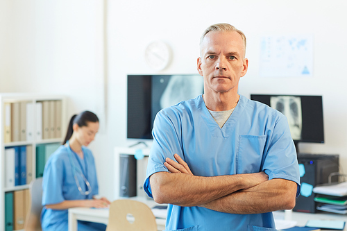 Waist up portrait of confident mature doctor  while standing with arms crossed in modern hospital interior, copy space