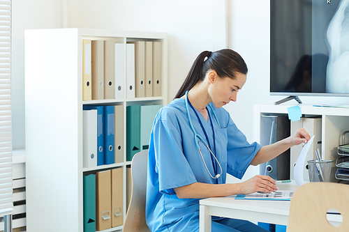 Side view portrait of female medic wearing blue uniform looking at clipboard while sitting at desk in clinic, copy space