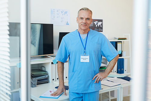 Waist up portrait of mature doctor wearing blue uniform smiling at camera while standing by desk in modern clinic, copy space