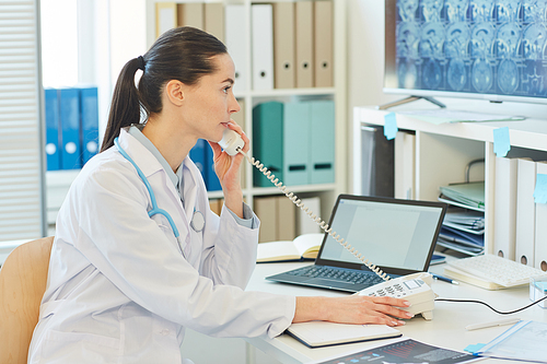 Side view portrait of young female doctor speaking by phone while sitting at desk and working in clinic or hospital, copy space