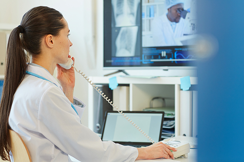 Side view portrait of female doctor speaking by corded phone while sitting at desk and working in clinic or hospital, copy space